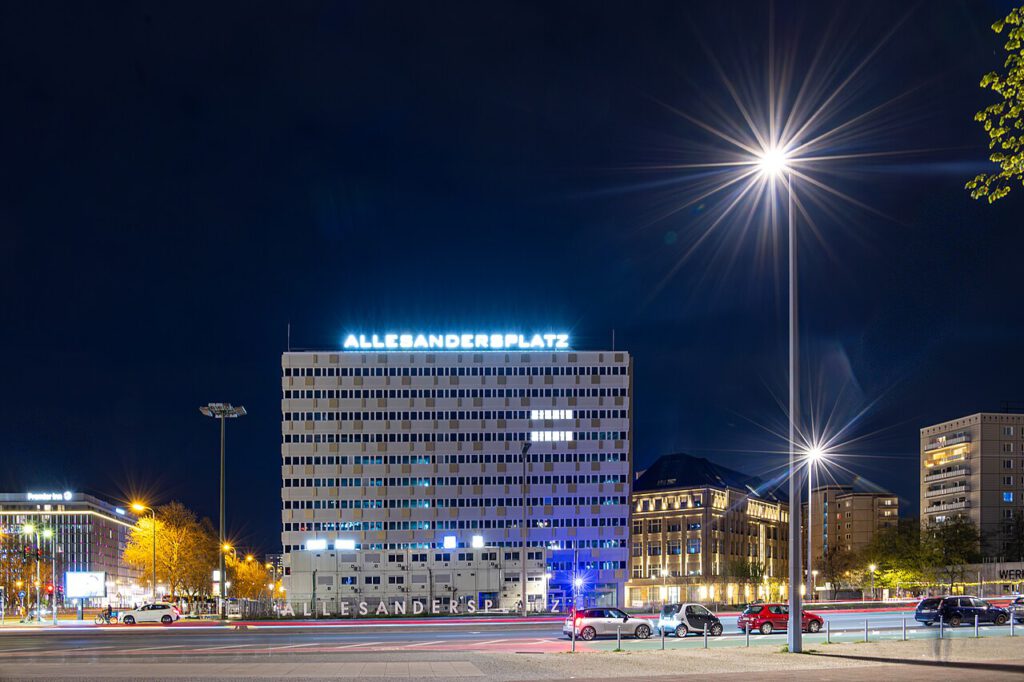 Nachtaufnahme des Allesandersplatz mit beleuchtetem Schriftzug, umgeben von modernen Gebäuden und Straßenbeleuchtung in einer urbanen Umgebung.