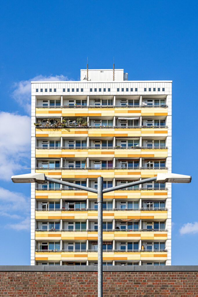Hochhaus mit gelben Balkonen und modernen Fenstern vor blauem Himmel, im Vordergrund eine Straßenlaterne.
