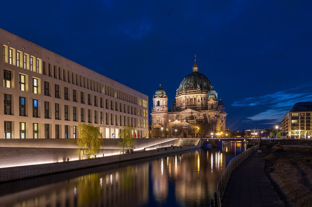 Nachtaufnahme des Berliner Doms und des Humboldt Forums, reflektiert im Wasser des umliegenden Kanals, mit beleuchteten Gebäuden und einem klaren, blauen Himmel.