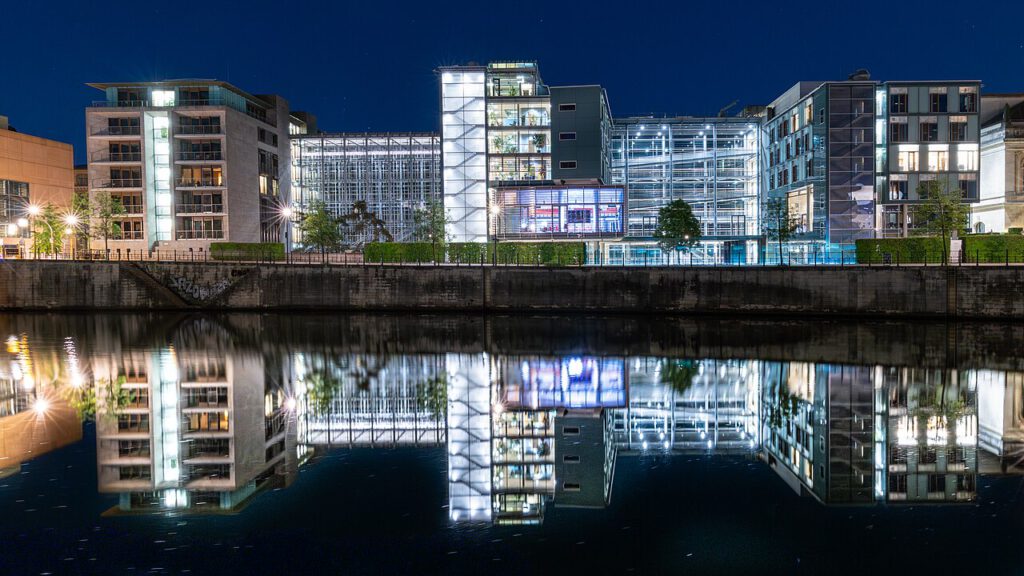 Moderne Architektur am Wasser bei Nacht mit reflektierenden Gebäuden und beleuchteten Fenstern. Die Umgebung zeigt eine harmonische Verbindung von Natur und urbanem Leben.