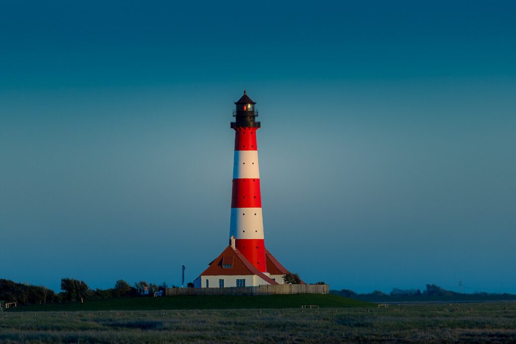 Leuchtturm mit rot-weißer Streifenmuster in einer ruhigen Landschaft, umgeben von Wiesen und Bäumen, bei klarem Himmel.