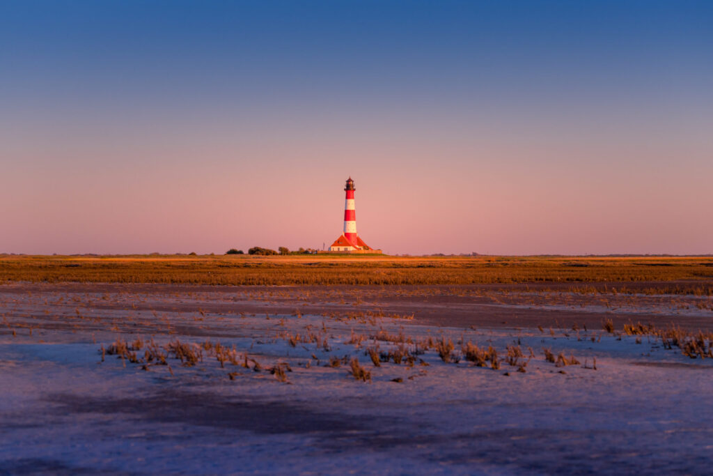 Leuchtturm im Wattenmeer bei Sonnenuntergang, umgeben von salzigen Wiesen und sanften Farben des Himmels, ideal für Küstenfotografie und Naturerlebnisse.