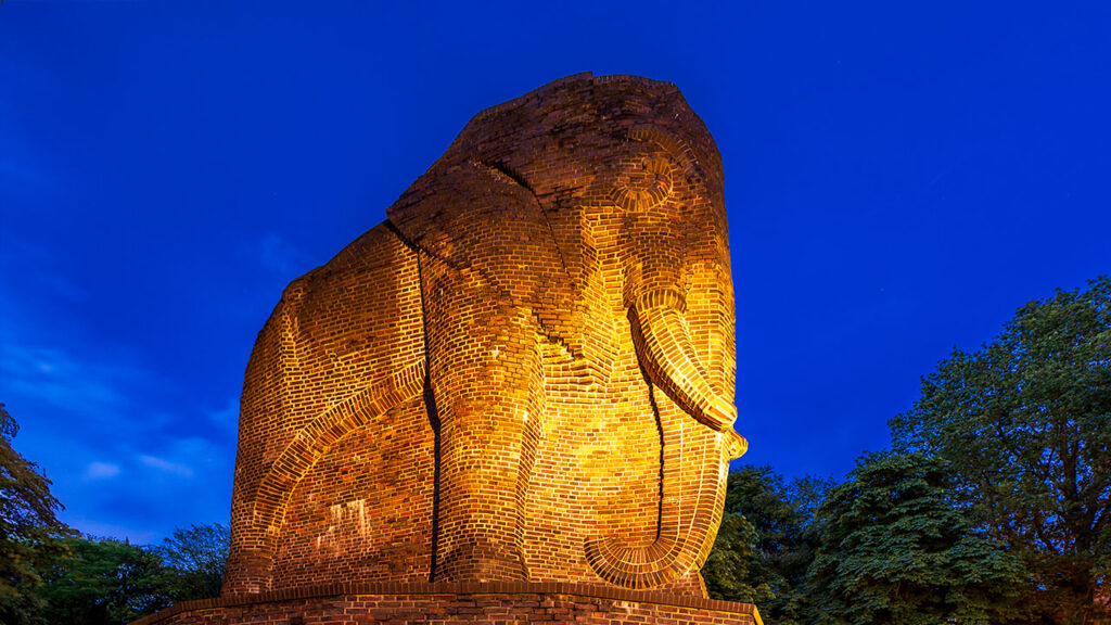 Das Antikolonialdenkmal in Bremen bei Nacht. Der aus Ziegelsteinen gemauerte Elefant wird angestrahlt und hebt sich so vom Hintergrund ab.