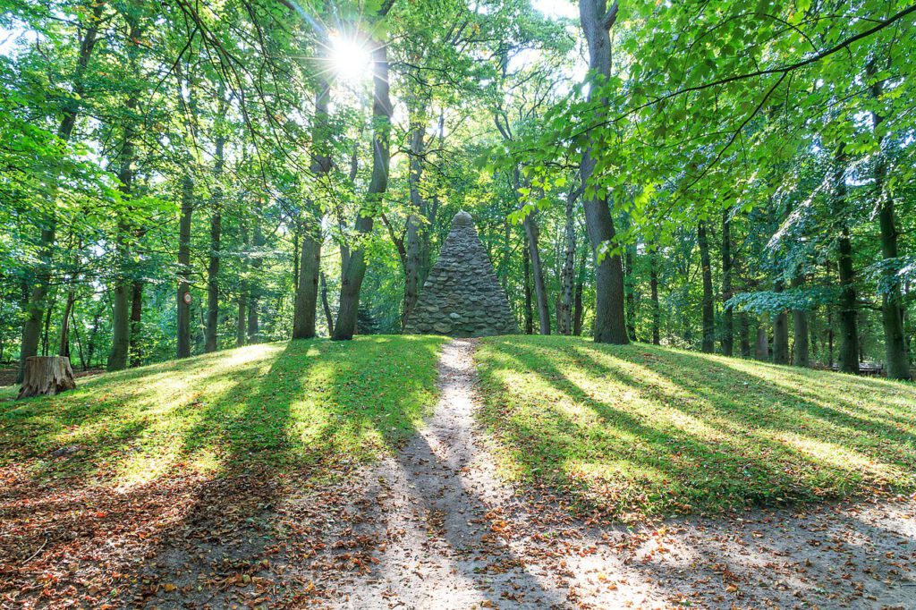 Das Upstalsboom-Denkmal, eine Pyramide im Wald, aufgenommen bei Gegenlicht. Sonnenlicht bricht durch das Laub und zeigt sich mit Strahlen.
