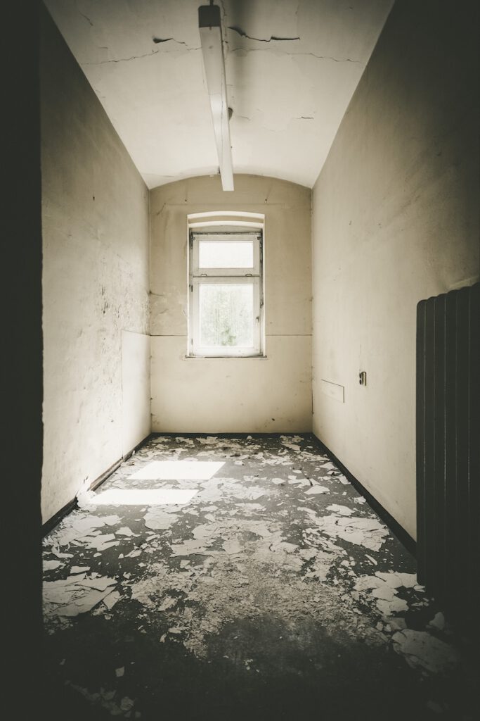 Blick in eine der Zellen im Erdgeschoss des Erweiterungsbaus von 1937. Sie sind eng und haben am Ende ein Fenster. Auf dem Boden liegt von der Wand oder Decke abgeblätterte Farbe.