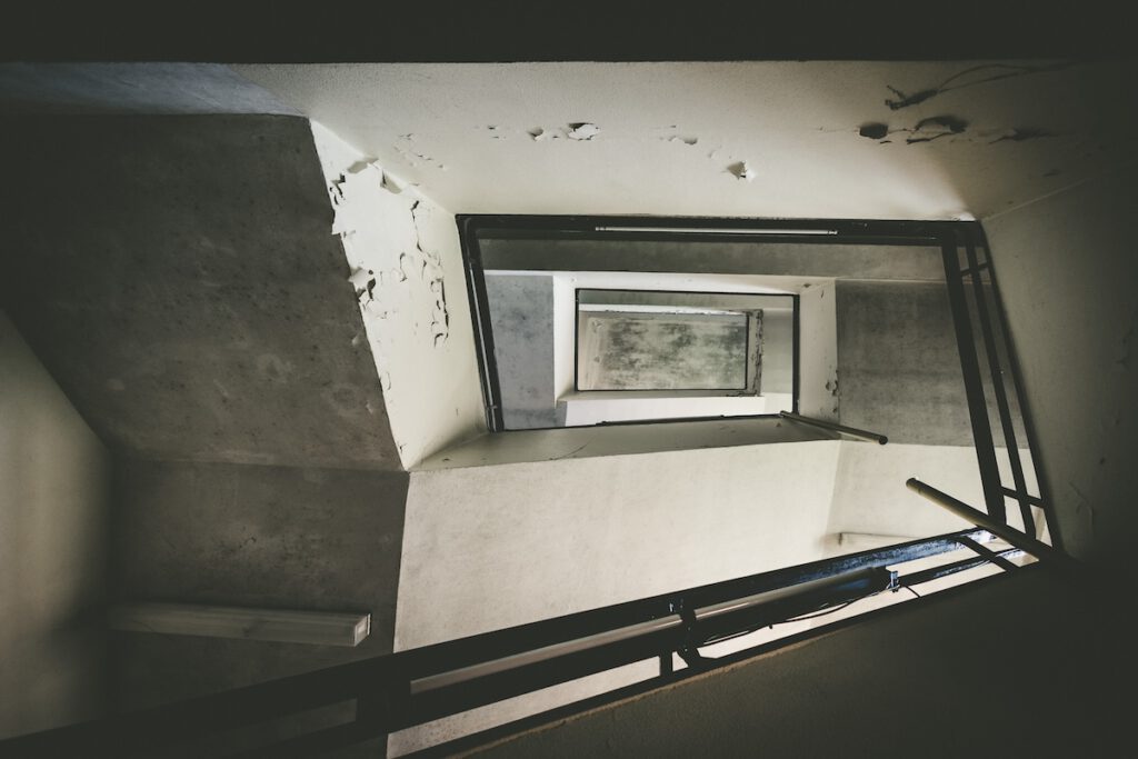 Blick in das Treppenhaus des Marineuntersuchungsgefängnis, von unten nach oben fotografiert. Es ist kantig und läuft um einen kleinen Innenraum herum. Überall blättert Farbe ab.