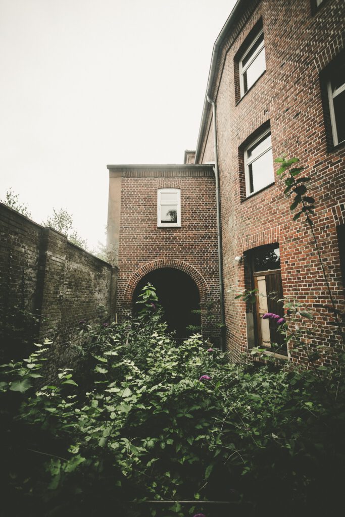 Blick in den heute mit Grünpflanzen überwucherten Gefängnishof. Im Hintergrund ist das Tor zu erkennen, über das LKW zugang zum Komplex hatten. Rechts die Außenmauer des Gefängnisgebäudes, linkes grenzt eine hohe Mauer das Marineuntersuchungsgefängnis vom Nachbargrundstück ab.