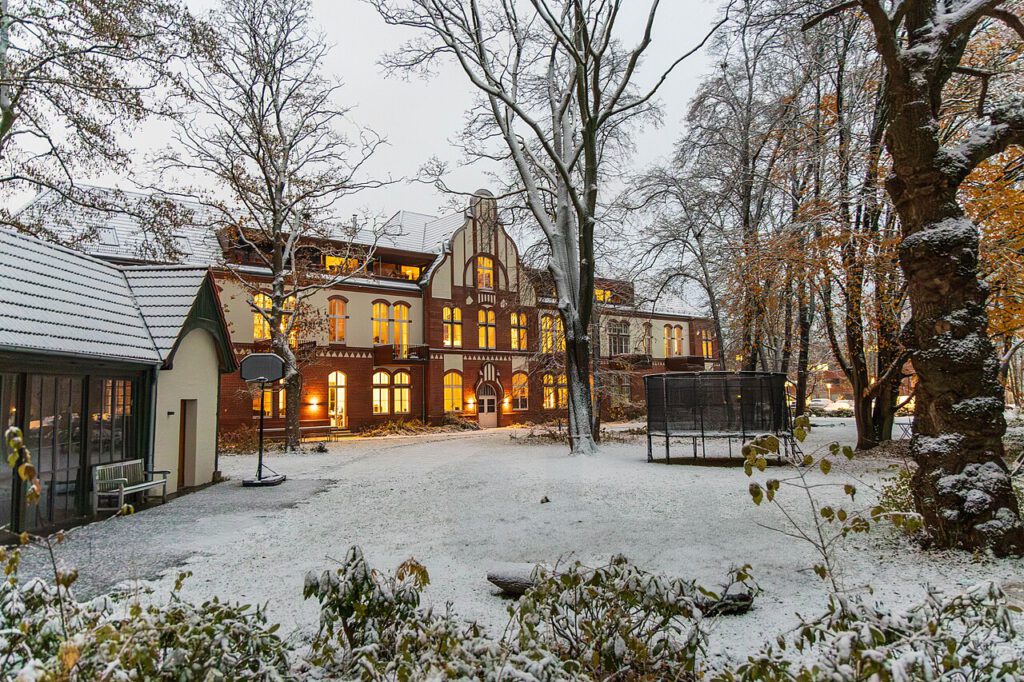 Blick auf das ehemalige Absonderungshaus des Krankenhauses. Es ist warm beleuchtet. Die Rasenflächen davor sind mit Schnee bedeckt. Das Gebäude dient heute als Wohngebäude