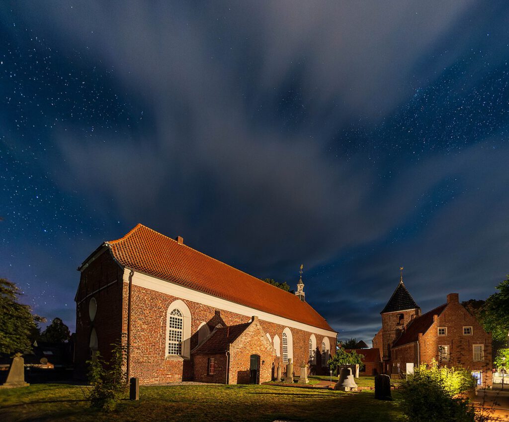 Nachtaufnahme von Kirche und Friedhof in Greetsiel. Die Backsteinkirche und der Friedhof werden vom Licht einer Laterne angeleuchtet, während weite Bereiche im Schatten versinken.