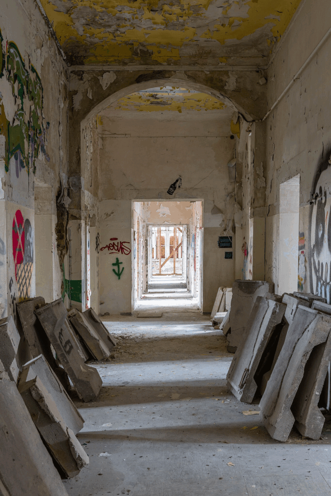 Aufnahme des verfallenen Flures im Erdgeschoss. Flure führen von ihm in die Krankenzimmer. An den Wänden stehen Betonplatten. Es sind Grafitti zu sehen. Der Putz ist teilweise abgeschlagen.