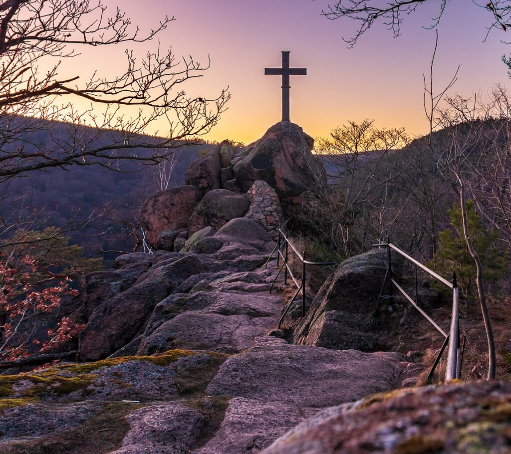 Der Ilsestein im Harz bei Sonnenuntergang, gekrönt von einem Gipfelkreuz. Die warmen Farben des Abendhimmels tauchen die markante Felsformation und die umliegende Landschaft in ein stimmungsvolles Licht.