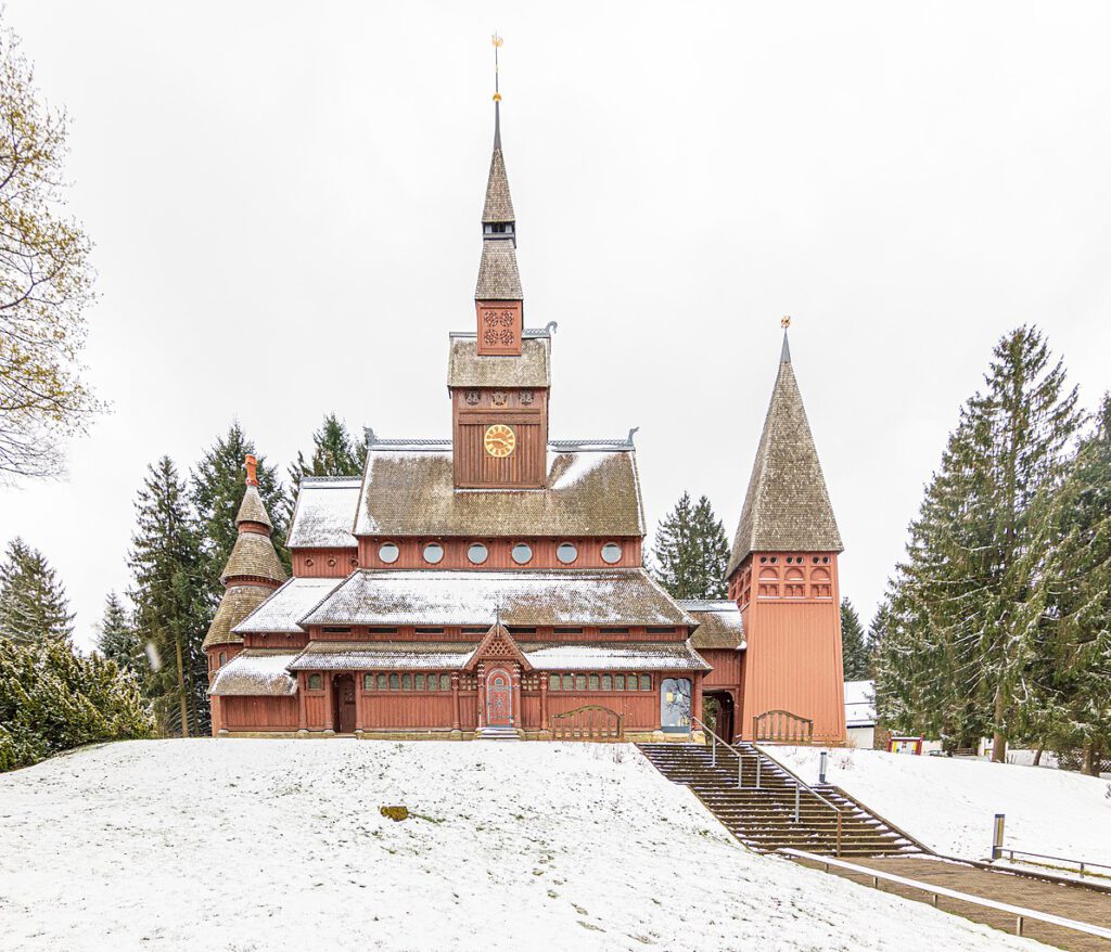 Die Stabkirche in Hahnenklee im Schnee, umgeben von einer winterlichen Landschaft. Die markante Architektur der Kirche, mit ihren spitzen Dächern und geschnitzten Holzdetails, hebt sich eindrucksvoll vor dem weißen Schneehintergrund ab