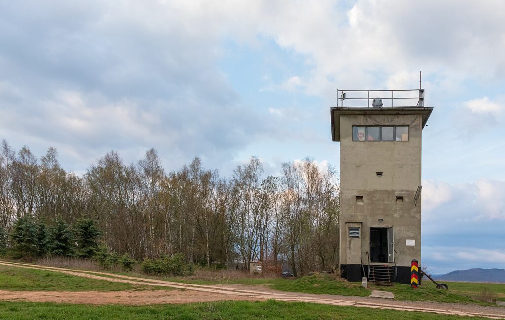 Ein gut erhaltener DDR-Grenzturm bei Bad Lauterberg, flankiert von einer markanten Grenzsäule mit den Farben Schwarz, Rot und Gold. Die Szene zeigt ein Stück deutscher Geschichte inmitten einer ländlichen Landschaft.