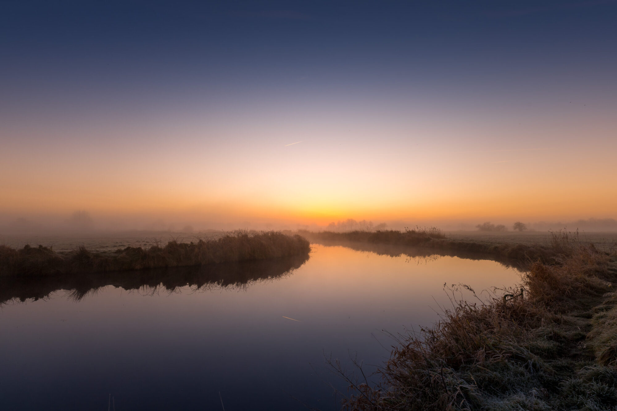 Ein ruhiger Fluss bei Sonnenaufgang, umgeben von einer nebligen Landschaft. Der Himmel zeigt einen sanften Übergang von tiefem Blau zu warmem Orange, das Wasser spiegelt die Farben wider. Die Ufer sind mit Gras und Schilf bewachsen, während die Ferne von dunstigen Silhouetten von Bäumen geprägt ist.