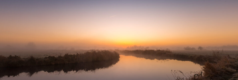 Ein ruhiger Fluss bei Sonnenaufgang, umgeben von einer nebligen Landschaft. Der Himmel zeigt einen sanften Übergang von tiefem Blau zu warmem Orange, das Wasser spiegelt die Farben wider. Die Ufer sind mit Gras und Schilf bewachsen, während die Ferne von dunstigen Silhouetten von Bäumen geprägt ist.