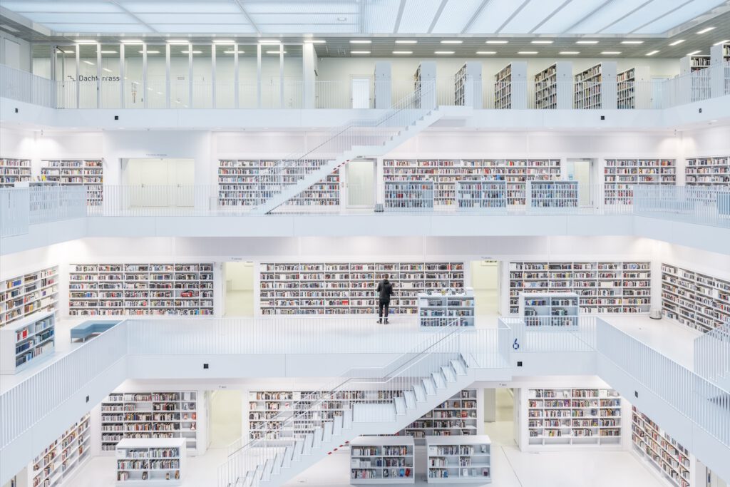 Innenansicht der Stadtbibliothek Stuttgart: Ein Mann steht im Zentrum der weißen, modernen Architektur mit treppenförmigen Ebenen und zahlreichen Bücherregalen. Mit dem Rücken zum Betrachter hält er ein Buch in der Hand und scheint vertieft zu lesen.