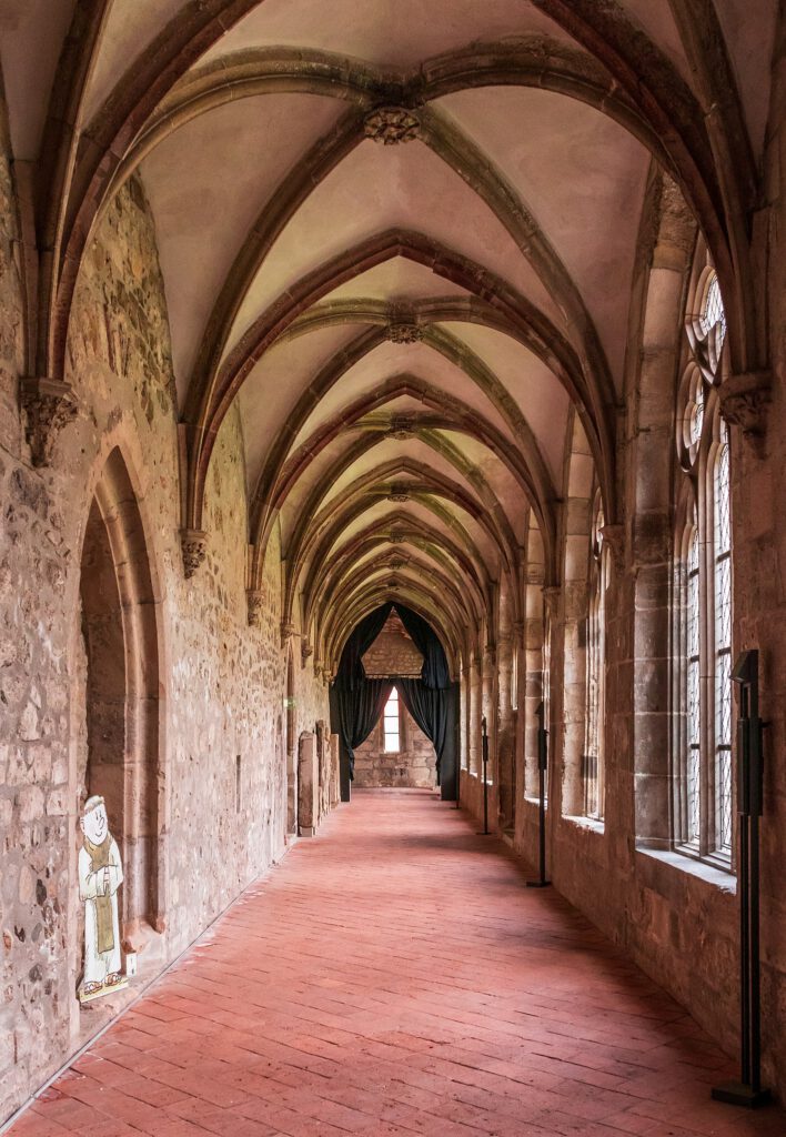 Ein Kreuzgang mit einem offenen, geschwungenen Vorhang am Ende, der den Blick auf eine Statue freigibt. Die Szene kombiniert die gotische Architektur mit einer geheimnisvollen und kunstvollen Inszenierung.