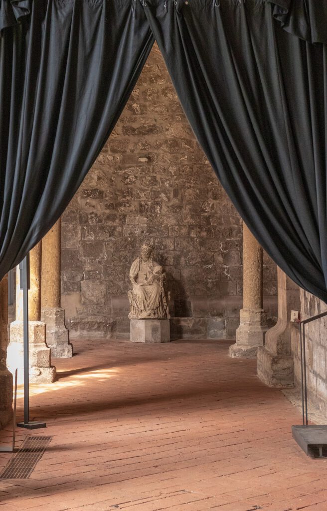 Eine gotische Madonna im Kreuzgang des Klosters Walkenried, elegant inszeniert hinter einem geöffneten Vorhang im Vordergrund. Die Statue steht im Zentrum der Aufmerksamkeit und wird von der historischen Architektur des Kreuzgangs umrahmt.