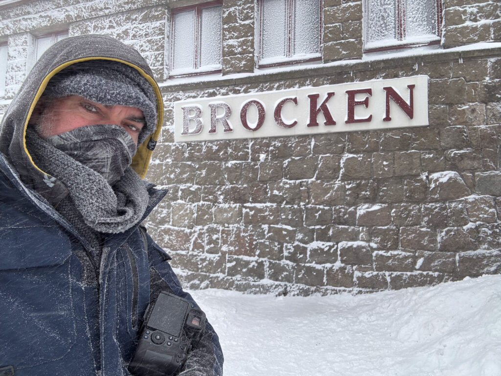 Matthias Süßen steht vor dem verschneiten Brockenbahnhof. Matthias ist dick in wintertaugliche Bekleidung gehüllt. Das Bild lässt erkennen, dass es sehr kalt ist und als Selfie mit dem Smartphone geschossen wurde.