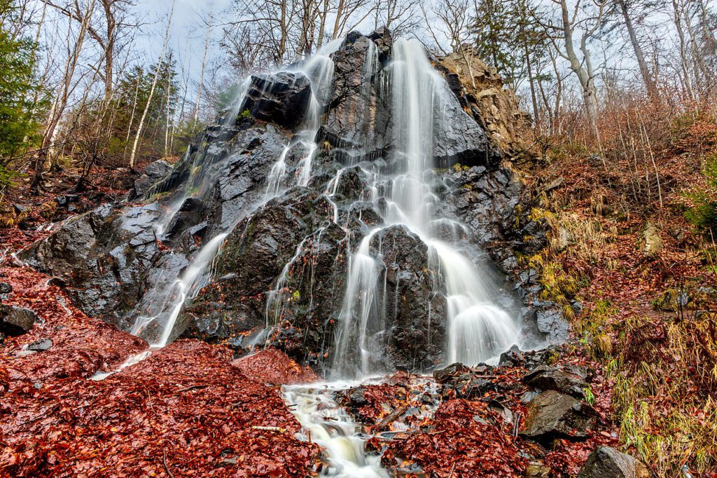 Der Radauwasserfall im Harz in einer Langzeitbelichtung. Das Wasser fließt in weichen, weißen Strömen über die Felsen, umgeben von herbstlichem, leuchtend rotem Laub, das die Szenerie in warme Farben taucht.