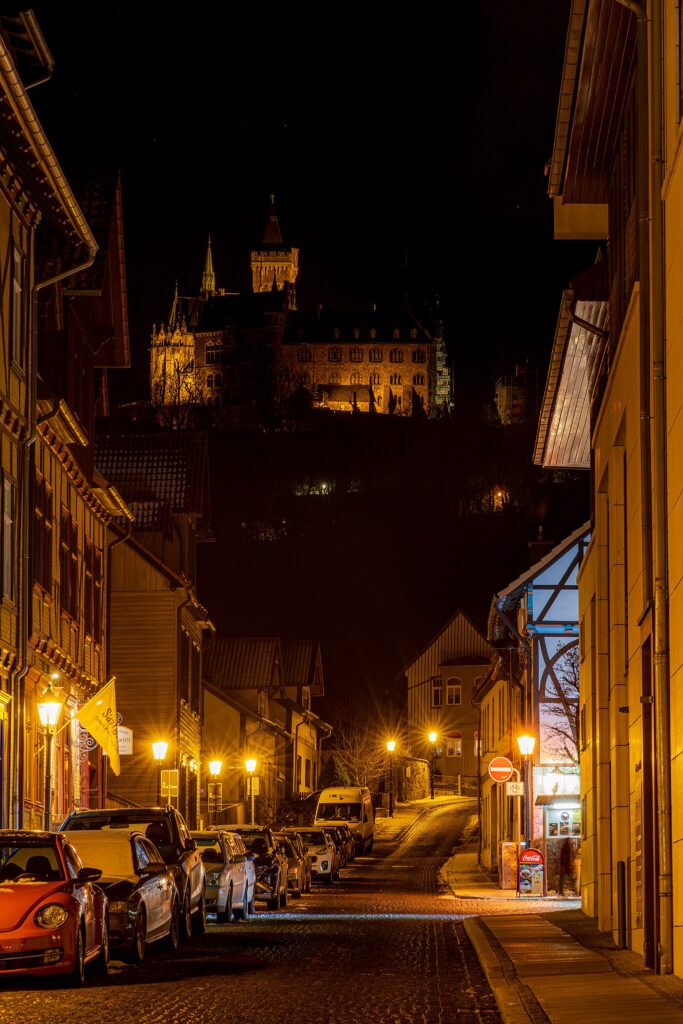 Nachtaufnahme einer schwach beleuchteten Straße in Wernigerode. Die sanfte Beleuchtung wirft ein gedämpftes Licht auf die Straße, während im Hintergrund hoch oben das imposante Schloss Wernigerode in der Dunkelheit sichtbar ist.