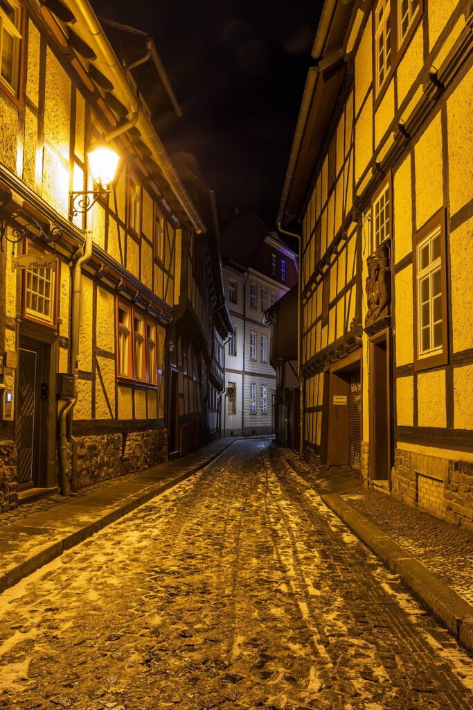 Nachtaufnahme einer menschenleeren, schwach beleuchteten Kopfsteinpflasterstraße in Wernigerode. Eine einzelne Straßenlaterne spendet schwaches Licht und wirft sanfte Schatten auf die Pflastersteine, was eine ruhige und geheimnisvolle Atmosphäre erzeugt.