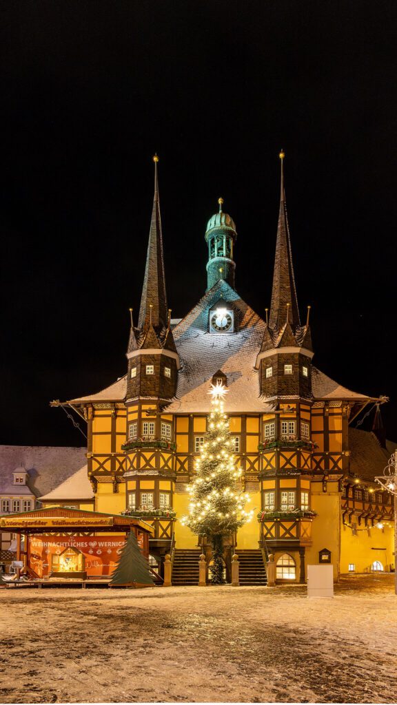 Nachtaufnahme des Rathauses von Wernigerode, festlich beleuchtet, mit einem prachtvollen Weihnachtsbaum davor. Die Lichter des Baums und die warme Beleuchtung des Rathauses schaffen eine festliche und einladende Atmosphäre.