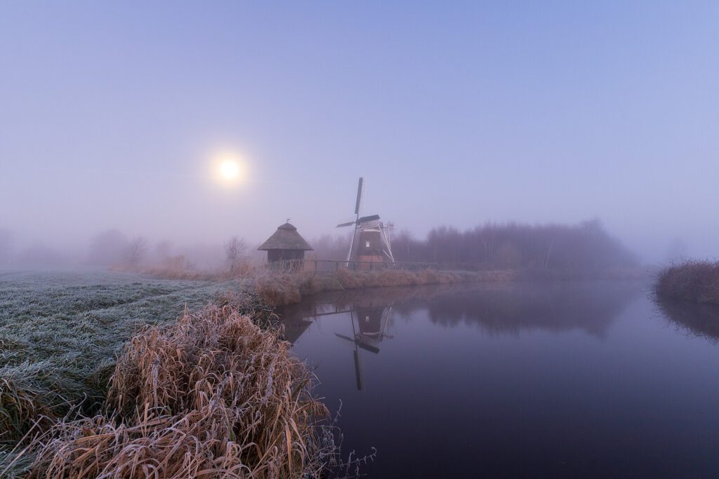 Eine idyllische Szenerie mit der historischen Windmühle Agnes  am Ufer eines ruhigen Flusses, umgeben von frostbedecktem Gras und Schilf. Der Himmel ist neblig-blau, der tiefstehende Mond scheint diffus durch den Dunst und beleuchtet die Szenerie spärlich. Die Windmühle und ein kleines, strohgedecktes Häuschen spiegeln sich im stillen Wasser wider, was der Szene eine verträumte Atmosphäre verleiht