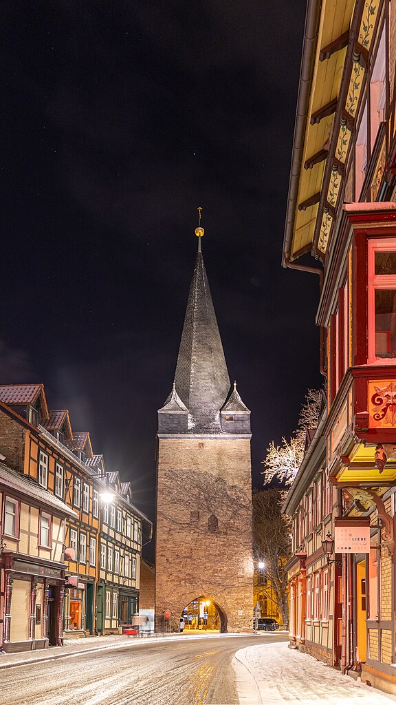 Nachtaufnahme des Westertores in Wernigerode, ohne Autos, im Schnee. Die historische Toranlage ist von einer dünnen Schneedecke bedeckt, und die sanfte Beleuchtung des Tors hebt die Details der Architektur hervor, während der Schnee eine ruhige und winterliche Stimmung verbreitet