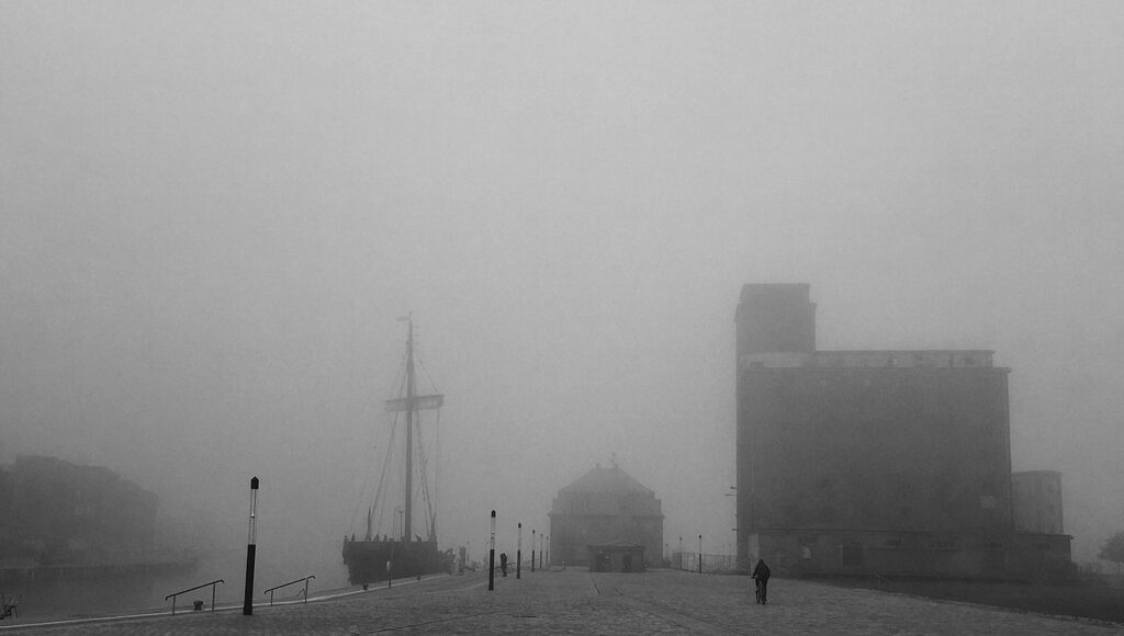 Der Hafen von Wismar in Schwarz-Weiß. Im Vordergrund führt ein gepflasterter Weg mit wenigen Passanten. Im Hintergrund sind die schemenhaften Konturen eines Segelschiffs, eines historischen Gebäudes und eines Silos zu erkennen, die im dichten Nebel fast verschwinden. Die Szenerie wirkt still und geheimnisvoll.