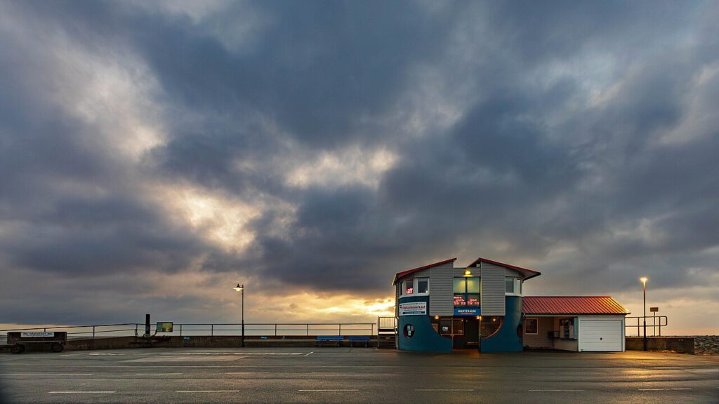 Wartehaus am Fährhafen Strucklahnungshörn auf Nordstrand bei dramatischer Wolkenstimmung. Die untergehende Sonne bricht durch die dunklen Wolken und taucht die Szene in ein stimmungsvolles Licht. Das markante Gebäude mit blauem Unterbau und rotem Dach hebt sich von der leeren, nassen Straße im Vordergrund ab.