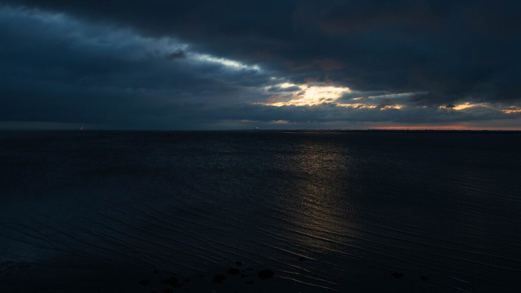 Abendstimmung am Fährhafen Strucklahnungshörn auf Nordstrand. Dunkle Wolken bedecken den Himmel, während ein schmaler Streifen Sonnenlicht durch die Wolkendecke bricht und sich auf der Wasseroberfläche spiegelt. Am Horizont sind die Lichter der Küste und eines Leuchtturms zu erkennen.