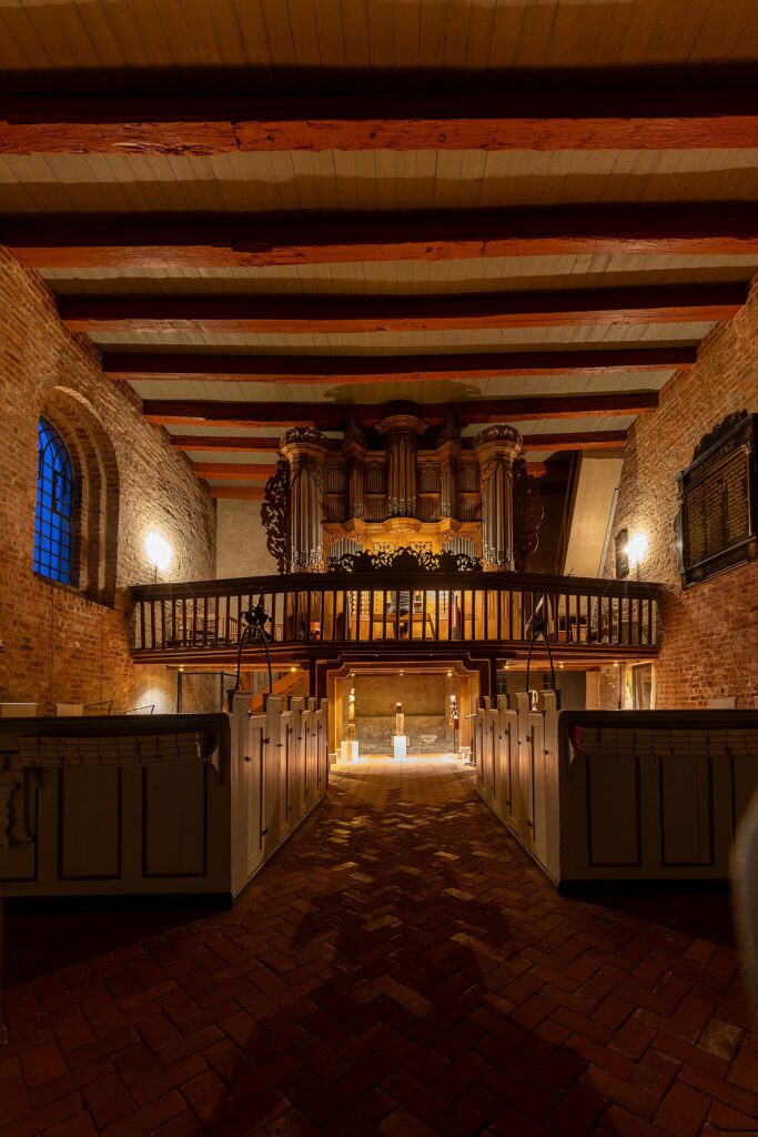 Innenraum der Alten Kirche auf Pellworm mit Blick auf die historische Orgel auf der hölzernen Empore. Die warm beleuchteten Backsteinwände und die hölzerne Balkendecke verleihen dem Raum eine gemütliche Atmosphäre. Im Vordergrund stehen schlichte graue Holzbänke, während im Hintergrund Skulpturen ausgestellt sind.