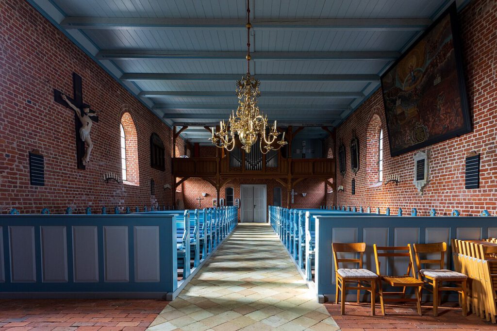 Innenraum der St. Crucis Kirche auf Pellworm. Die Kirche mit Backsteinmauern, blauen Holzbänken und einer hellblauen Holzdecke wird von einem goldenen Kronleuchter in der Mitte erleuchtet. An der linken Wand hängt ein großes Kruzifix, während an der rechten Wand ein historisches Gemälde zu sehen ist. Im Hintergrund befindet sich eine Orgel auf einer hölzernen Empore.