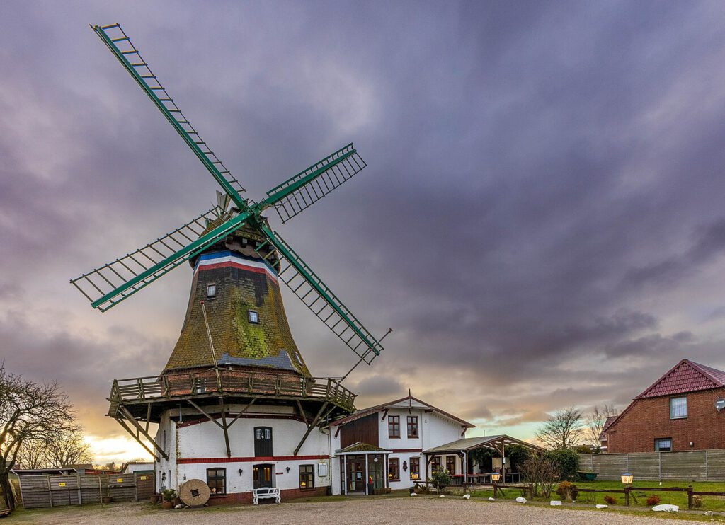 Alt-Text: Historische Windmühle Engel auf Nordstrand vor einem dramatischen, bewölkten Himmel. Die grün-weißen Flügel der Mühle erstrecken sich über das Bild, während das mit Moos bewachsene Mühlengebäude und angrenzende Gebäude eine ländliche, einladende Atmosphäre schaffen.