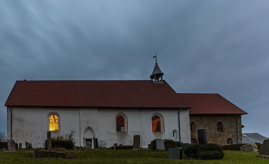 Außenansicht der alten Inselkirche Pellworm in der Dämmerung. Das Gebäude mit weiß verputzten Wänden und einem roten Ziegeldach steht auf einem Friedhof mit alten Grabsteinen. Durch die erleuchteten Fenster strahlt warmes Licht nach außen. Der Himmel ist von dichten Wolken bedeckt, was die Atmosphäre verstärkt.