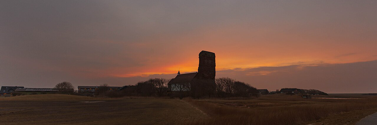 Silhouette der Alten Kirche St. Salvator auf Pellworm mit ihrer markanten Turmruine vor einem leuchtenden Sonnenaufgang. Der Himmel zeigt warme Orange- und Gelbtöne, während dunkle Bäume und Gebäude die Szenerie einrahmen.