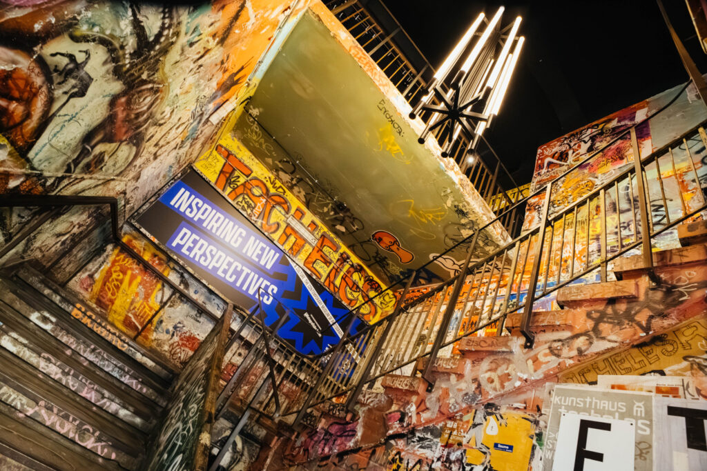 Bunt besprühtes Treppenhaus im ehemaligen Kunsthaus Tacheles in Berlin, heute Teil des Fotografiska Berlin. Die Wände und Stufen sind mit Graffiti und Streetart bedeckt, über dem Treppenaufgang hängt ein modernes Leuchtelement. Ein großes Schild mit der Aufschrift „Inspiring New Perspectives“ verweist auf die heutige Nutzung als Kunsthalle.