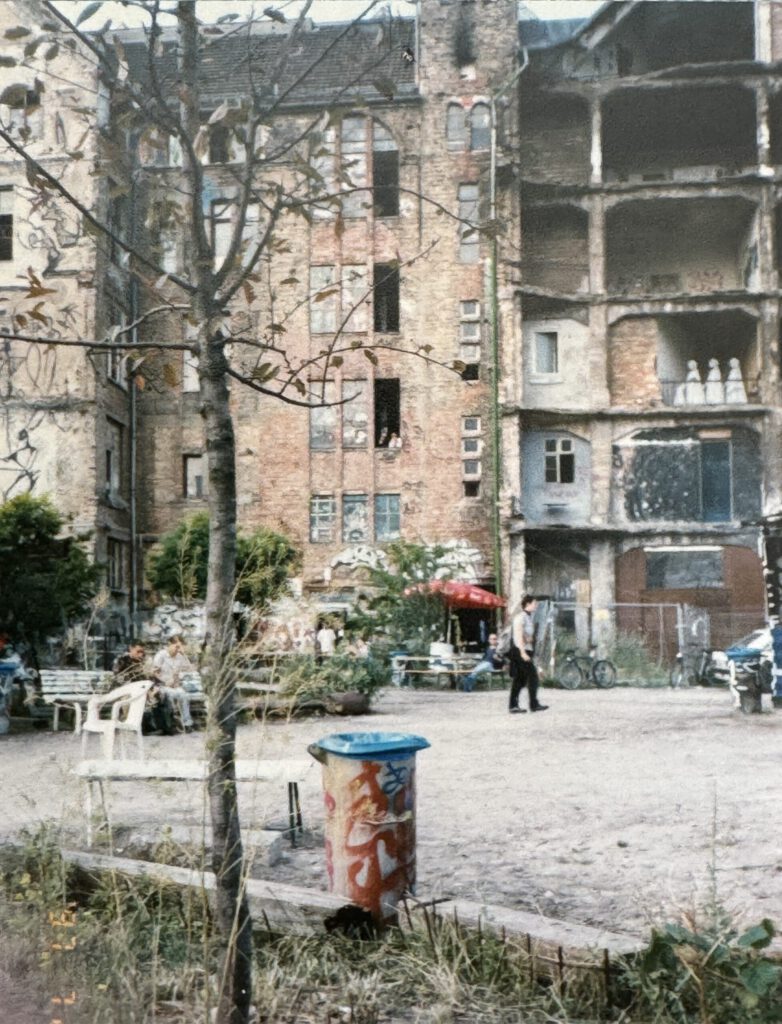 Innenhof des Kunsthauses Tacheles in Berlin, fotografiert im Jahr 1997. Im Vordergrund ein bunt besprühter Mülleimer, dahinter verstreute Bänke und Tische, an denen Menschen sitzen. Im Hintergrund das teils verfallene Hauptgebäude mit offenen Fassaden, Graffiti und improvisierter Begrünung. Ein Mann geht über den Hof, hinter einem roten Sonnenschirm befindet sich das damalige Café Zapata.