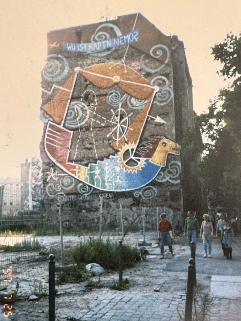 Wandbild am ehemaligen Kunsthaus Tacheles in Berlin, aufgenommen 1997. Das farbenfrohe Mural zeigt ein stilisiertes, fliegendes Schiff mit dem Schriftzug „Wo ist Käpt’n Nemo?“ an einer Feuermauer. Im Vordergrund ein brachliegendes Gelände, dahinter Passant:innen auf dem Gehweg in der Oranienburger Straße. Das Bild vermittelt den rauen Charme der Berliner 90er-Jahre-Kunstszene.