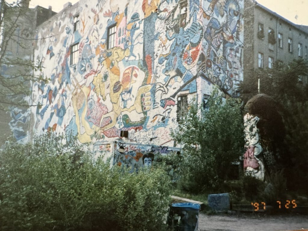 Rückseite des Kunsthauses Tacheles in Berlin, fotografiert am 25. Juli 1997. Die Hauswand ist großflächig mit einem farbenfrohen, detailreichen Wandbild bedeckt, das zahlreiche surrealistische Figuren und Symbole zeigt. Im Vordergrund überwucherter Garten, ein besprühter Mülleimer und Graffiti auf weiteren Wandflächen. Die Szene dokumentiert die künstlerische Ausdruckskraft der Berliner Subkulturszene der 90er-Jahre.