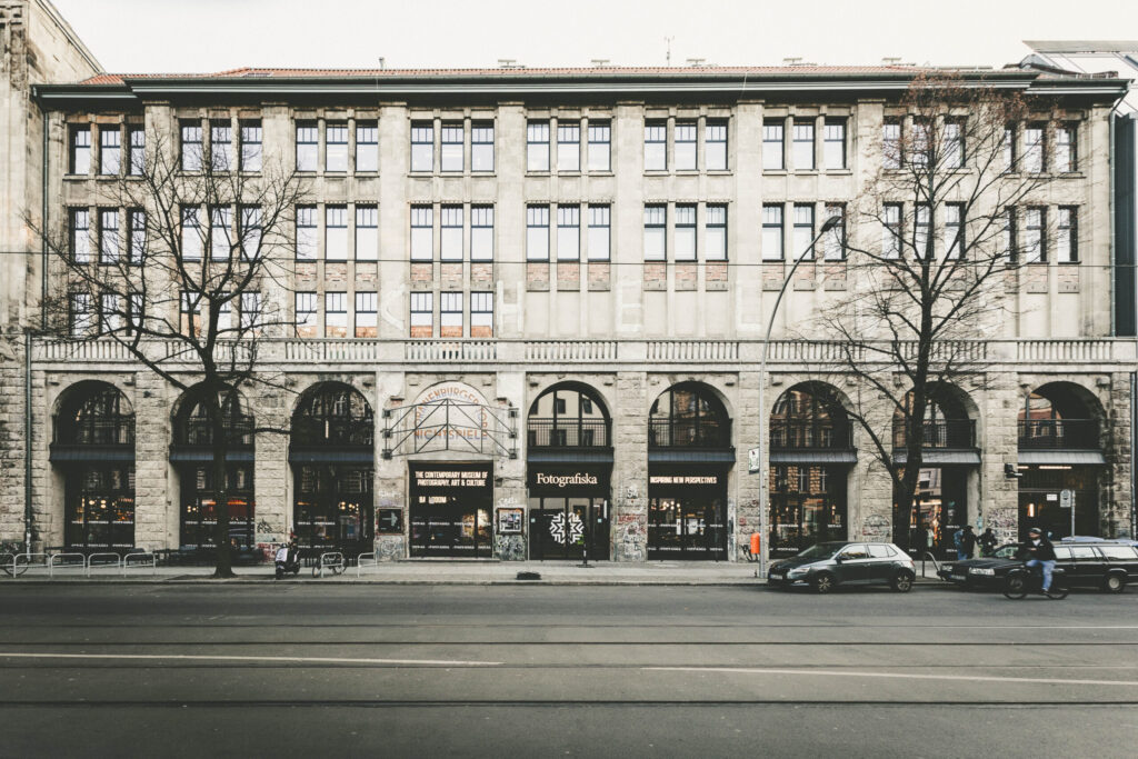 Frontansicht des Gebäudes der Fotografiska Berlin, einem Museum für zeitgenössische Fotografie, Kunst und Kultur. Das Gebäude ist mehrstöckig mit großen Fenstern und Rundbögen im Erdgeschoss. Über dem Eingang befindet sich ein Schriftzug mit dem Namen des ehemaligen Kunsthauses „Tacheles“. Vor dem Gebäude stehen Autos, Fahrräder und einige Passanten. Die Szene wirkt urban und leicht winterlich, mit kahlen Bäumen und gedämpften Farben.