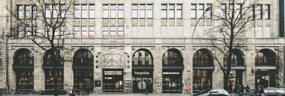 Frontansicht des Gebäudes der Fotografiska Berlin, einem Museum für zeitgenössische Fotografie, Kunst und Kultur. Das Gebäude ist mehrstöckig mit großen Fenstern und Rundbögen im Erdgeschoss. Über dem Eingang befindet sich ein Schriftzug mit dem Namen des ehemaligen Kunsthauses „Tacheles“. Vor dem Gebäude stehen Autos, Fahrräder und einige Passanten. Die Szene wirkt urban und leicht winterlich, mit kahlen Bäumen und gedämpften Farben.