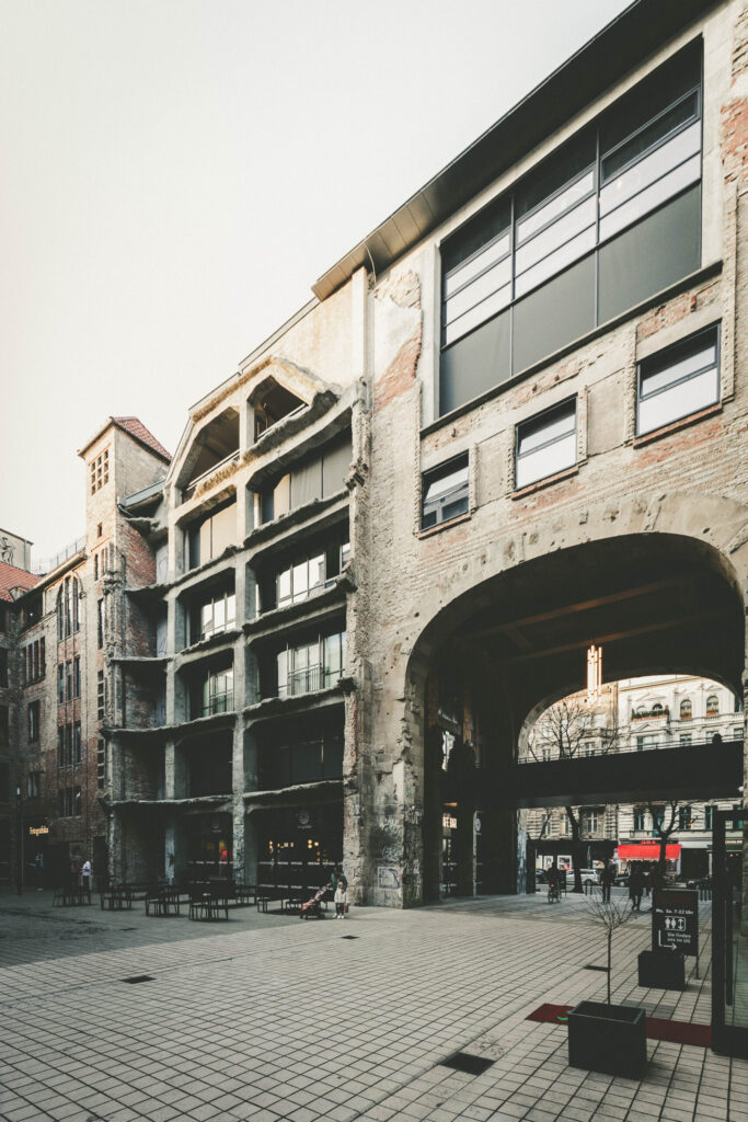 Aufnahme der sanierten Fassade des ehemaligen Kunsthauses Tacheles in Berlin im Jahr 2025. Die Struktur des alten Gebäudes ist noch deutlich zu erkennen, wurde jedoch modernisiert und mit großen Glasflächen versehen. Im Erdgeschoss befinden sich Cafés und Geschäfte, im Hintergrund ist der Eingang zum Fotografiska Berlin unter einem großen Rundbogen sichtbar. Die Umgebung wirkt aufgeräumt und architektonisch durchgestaltet.