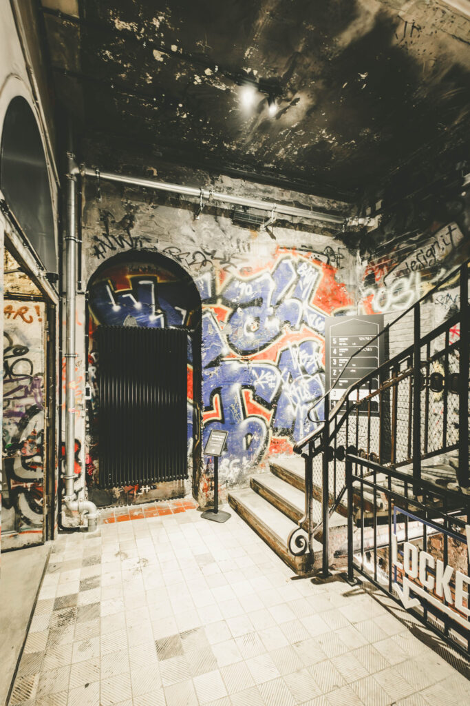 Eingangsbereich des ehemaligen Kunsthauses Tacheles in Berlin. Eine abgenutzte Steintreppe führt vorbei an einer mit farbenfrohem Graffiti bedeckten Wand und einem schwarzen Rundbogen. Die Decke ist dunkel und rußgeschwärzt, Metallrohre verlaufen offen entlang der Wände. Rechts zeigt ein modernes Schild mit weißen Lettern auf schwarzem Hintergrund die Etagenbelegung des Gebäudes. Am Geländer ist das Wort „Lockers“ mit einem Richtungspfeil angebracht. Der Kontrast zwischen Street Art, urbanem Verfall und strukturierter Besucherführung vermittelt den besonderen Charme dieses Ortes.
