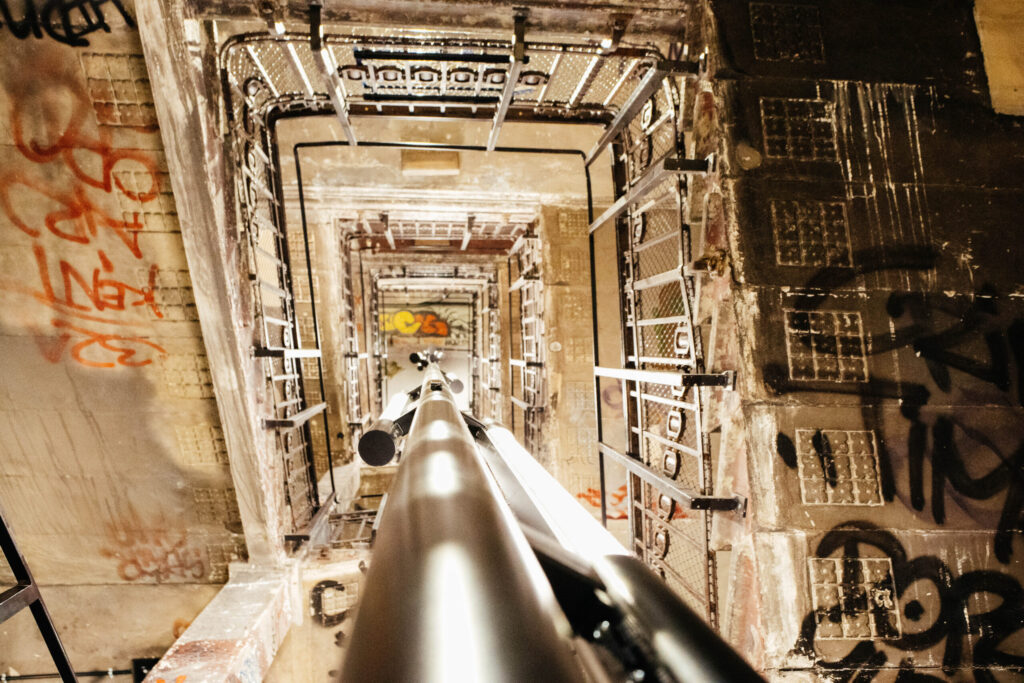 Blick nach oben durch das zentrale Treppenhaus des ehemaligen Kunsthauses Tacheles in Berlin. Die Perspektive zeigt ein scheinbar endloses Rechteck aus Metalltreppen, Geländern und Plattformen, alle dicht mit Graffiti bedeckt. Ein markanter Lichtstab verläuft vertikal durch die Bildmitte und verstärkt den Tiefeneffekt. Die Wände sind abgenutzt und mit Schriftzügen, Symbolen und Farbschichten bedeckt – ein Sinnbild für die alternative Kunstszene und die bewegte Geschichte des Gebäudes.
