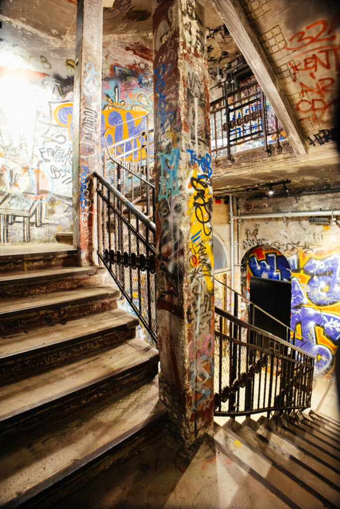Innenansicht eines mehrstöckigen Treppenhauses im ehemaligen Kunsthaus Tacheles in Berlin. Die Wände, Pfeiler und Treppenstufen sind vollständig mit bunten Graffiti, Tags und Farbschichten bedeckt. Die Metallgeländer mit Gitterelementen unterstreichen die rohe, industrielle Atmosphäre des Ortes. Die Beleuchtung wirft warme, diffuse Lichtakzente auf die verwitterten Oberflächen. Der Blick folgt den Treppenläufen nach oben und unten, wodurch ein labyrinthischer Eindruck entsteht – Symbol für die Vielschichtigkeit und kreative Energie des einstigen Künstlerkollektivs.