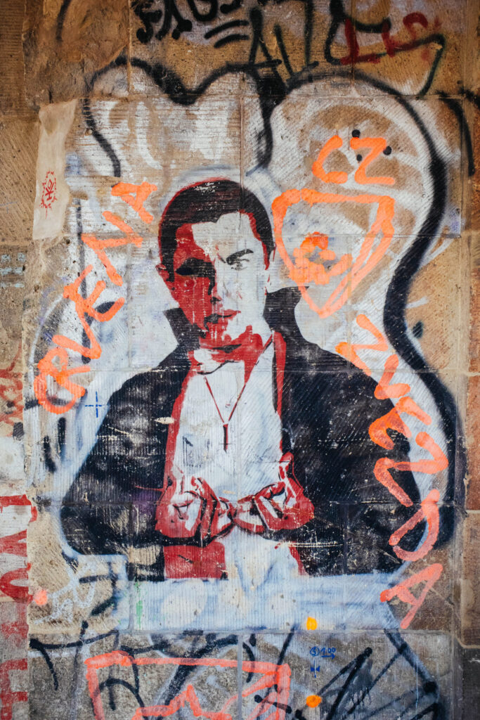 Streetart-Porträt eines Mannes im Stil von Dracula, in Schwarz, Weiß und Rot auf eine raue Steinwand gemalt. Die Figur trägt ein Medaillon und ein klassisches Vampirkostüm mit Umhang. Um das Bild herum sind orangefarbene und schwarze Graffiti und Tags zu sehen, darunter stilisierte Schriftzüge und Herzformen. Das Kunstwerk wirkt geheimnisvoll und theatralisch, ergänzt durch die kontrastreiche Umgebung – typisch für die rebellische, collageartige Ästhetik des ehemaligen Berliner Kunsthauses Tacheles.