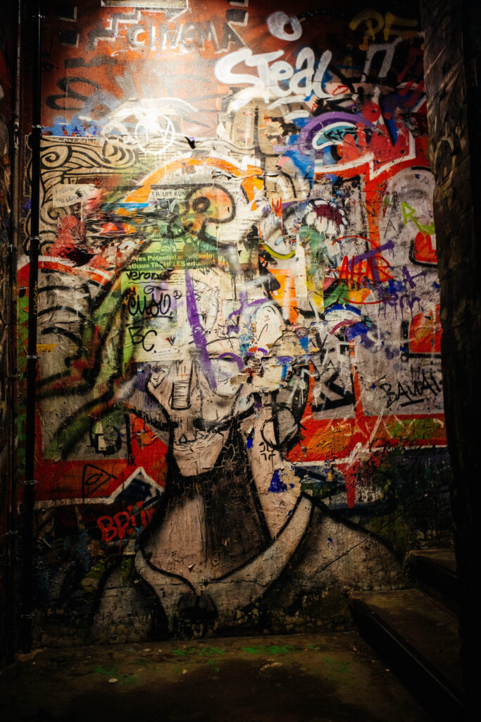 Bunte, chaotische Wand im ehemaligen Kunsthaus Tacheles, überzogen mit zahllosen Graffiti, Farbschichten und Kritzeleien. Im unteren Bereich ist ein verblasstes Wandbild eines Mannes mit Brille und Zigarre zu erkennen, das teilweise übermalt wurde. Der Hintergrund ist dicht überlagert mit Schriftzügen, Symbolen und Collage-Fragmenten in leuchtenden Farben. Der Boden ist abgenutzt, der Raum wirkt roh, authentisch und rebellisch – Sinnbild für die subkulturelle Energie des Ortes.