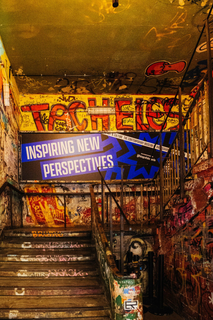 Blick auf ein mit Graffiti übersätes Treppenhaus im ehemaligen Kunsthaus Tacheles in Berlin. An der Wand prangt in großen, bunten Buchstaben das Wort „TACHELES“. Darunter hängt ein modernes Werbebanner des Fotografiska Museums mit dem Slogan „Inspiring New Perspectives“. Die Wände, Geländer und Stufen sind dicht mit Schriftzügen, Stickern und Farbspuren bedeckt. Die Szene kombiniert den rauen, rebellischen Charakter der Tacheles-Vergangenheit mit dem zeitgenössischen Auftritt der heutigen Museumseinrichtung.